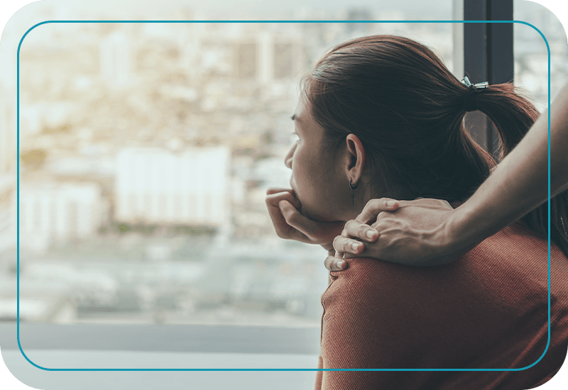 Image of woman staring sadly out window, someone placing their hand on her shoulder